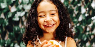 A young child laughing joyfully outdoors in front of a leafy wall, holding an apple or snack, symbolizing happy childhood, emotional well-being, and positive parenting.