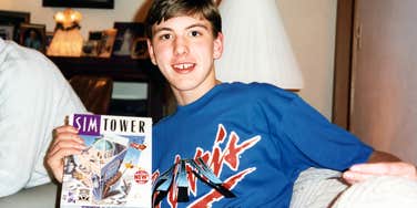 A teenage boy sitting on a couch holding a Maxis PC game box in a 1990s living room setting, symbolizing 90s nostalgia, PC gaming, and retro childhood.