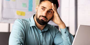 A close-up portrait of a thoughtful Hispanic businessman in a light blue shirt resting his forehead on his hand while looking away from his laptop.