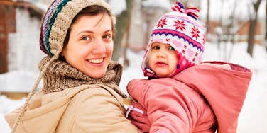 A cheerful mother wearing a knitted beanie holding her toddler warmly bundled in a pink winter coat outdoors in the snow.