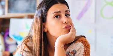 A portrait of a young female teacher in a knit sweater leaning her chin on her hand with an exasperated face in a classroom.