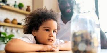 Child gazes at a jar of coins while a parent looks on, the small moments money talk shapes.
