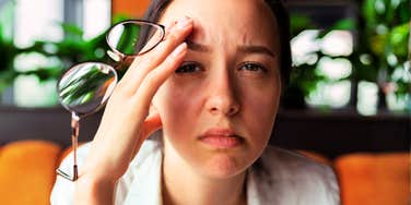 A young woman touching her head with a weary, stressed expression while holding her eyeglasses.
