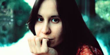 A close-up portrait of a young brunette woman resting her chin on her hand, looking directly at the camera with a contemplative expression.