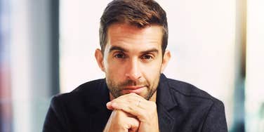 A close-up portrait of a thoughtful businessman resting his chin on his clasped hands in a modern office environment.
