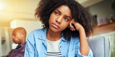 Woman looks away, troubled, while her partner sits distant behind her, the quiet strain toxic words can leave.
