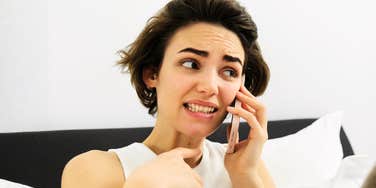 A close-up portrait of a young woman with short dark hair making an awkward grimace while talking on her smartphone in bed.