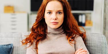 A red-haired woman looking steadily into the camera with a serious facial expression while crossing her arms on a sofa, symbolizing high standards, skepticism, and analytical focus.