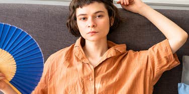 woman with zero energy looks sternly into camera holding a paper fan