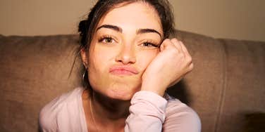 A young woman sitting on a couch in warm indoor lighting looking exasperated while pouting, symbolizing generational miscommunication, annoyance, and workplace culture dynamics.