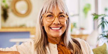 A close-up portrait of a cheerful senior woman with grey hair and glasses smiling warmly at home.