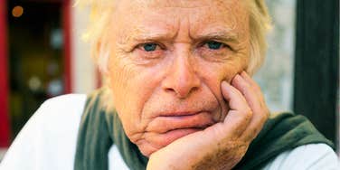 An elderly man with blue eyes and light blonde hair looking directly at the camera with a pensive expression while leaning his face on his hand, symbolizing family strain, grief, and reflection.