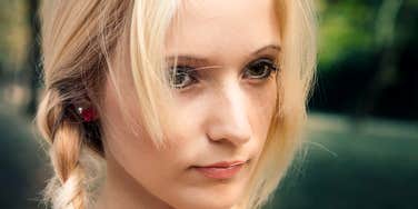 A close-up portrait of a young blonde woman with braided hair looking down and to the side with a serious, pensive expression.