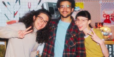 A group of three smiling teenagers in 90s retro clothing posing together cheerfully with a boombox.