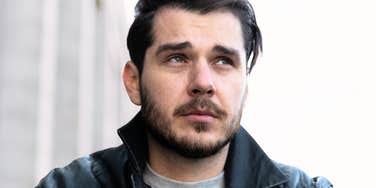 A dark-haired man with a short beard and leather jacket sitting outside, looking thoughtfully up into the distance with a serious expression, representing self-reflection, late-diagnosed autism, and processing internal fatigue.