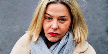 A close-up portrait of a middle-aged woman with blonde hair, red lipstick, and a gray scarf over a beige fleece coat, gazing with a tired, solemn expression, representing parental heartbreak, acceptance, and quiet resilience.