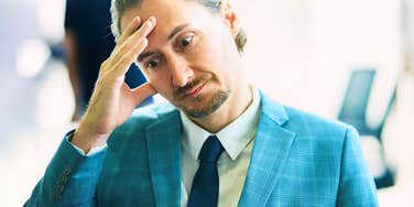 A medium close-up portrait of a businessman in a light blue patterned suit and dark blue tie holding his hand to his forehead with a stressed, regretful expression, representing recruitment failure, business stress, and management missteps.