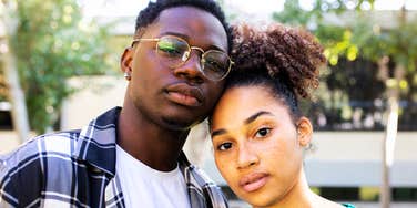 A young Black couple standing outside, leaning their heads together while staring directly into the lens with solemn, unsmiling eyes, representing relationship facade, emotional disconnection, and hidden marital strain.