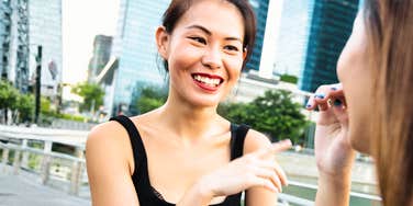 A bright photo of an Asian woman in a black tank top smiling warmly and talking with a friend outdoors near a city waterfront, representing casual charisma, effortless connection, and engaging social conversation.