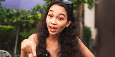 Entitled woman up close with dark curly hair and sunglasses on top of her head, pointing her finger assertively downward while speaking with a surprised demanding facial expression. 