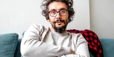 A close-up portrait of a middle-aged man with curly dark hair, glasses, and a beard sitting on a teal couch with his arms tightly crossed, gazing at the camera with a stubborn, distant expression, representing emotional sulking and defiance.