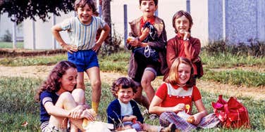 Group of six smiling children from the 1970s sitting and standing on a grassy field, eating snacks.