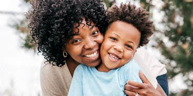 A smiling Black mother with curly hair hugging her cheerful young son outdoors, both looking at the camera with genuine joy.