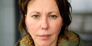 Woman with dark brown hair and hazel eyes looking directly at the camera with a calm, serious expression, wearing an olive green jacket with a shearling collar.