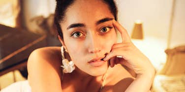 Woman with dark hair pulled back resting her chin on her hand and looking directly at the camera with a thoughtful expression in warm light.