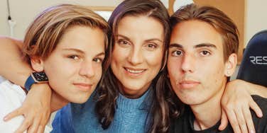 A close-up portrait of a smiling mother in a blue sweater hugging her two teenage sons closely, with both boys leaning in warmly toward the camera, representing affectionate parenting, strong family bonds, and emotional warmth.