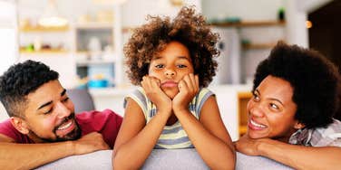 A young Black girl sitting between her smiling mother and father on a sofa with a sad, overwhelmed expression resting her head on her hands.