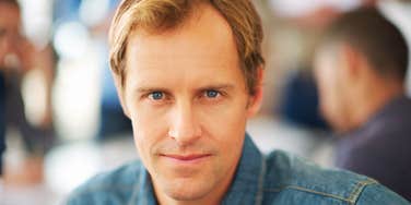 Man with light brown hair and blue eyes wearing a denim shirt, looking at the camera with a subtle, knowing smirk in a blurred café setting.