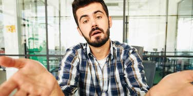 A close-up portrait of a dark-haired man in a blue and white plaid shirt sitting in an office, gesturing with open hands and a skeptical, dismissive expression toward the camera, representing passive-aggressive communication and defensiveness.