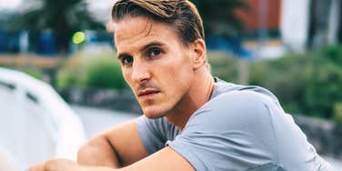 A close-up profile portrait of a handsome blonde man in a grey athletic t-shirt leaning against a railing outdoors, looking at the camera with a somber, serious, and detached expression, representing emotional distance and relationship tension.