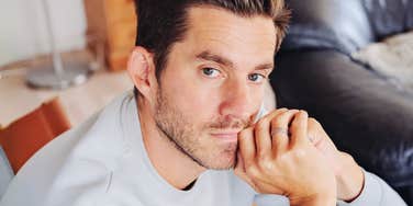 A close-up portrait of a brown-haired man in a light grey sweater resting his chin on his clasped hands, prominent wedding ring visible, looking directly at the camera with a somber, unsmiling, and weary expression indoors.