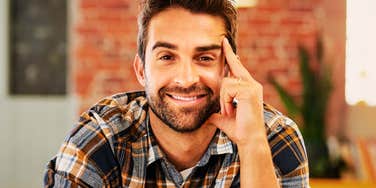 A close-up portrait of a young man with a goatee and thin-framed glasses wearing a burgundy sweater, looking away with a neutral, detached expression in soft sunlight, representing emotional distance, indifference, and introspection.