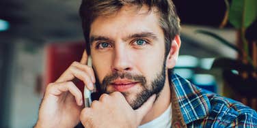 Bearded man in a plaid shirt holding a phone to his ear with his other hand at his chin, looking directly at the camera with a thoughtful expression.