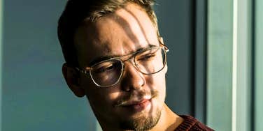 A close-up portrait of a young man with a goatee and thin-framed glasses wearing a burgundy sweater, looking away with a neutral, detached expression in soft sunlight, representing emotional distance, indifference, and introspection.