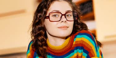 Brunette woman with curly hair wearing a colorful stripe sweater, looking at camera with a curious smile.