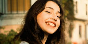 A close-up portrait of a young woman with long dark hair smiling broadly with closed eyes in warm golden hour sunlight, wearing a light denim jacket, representing optimism, joy, and open-mindedness.