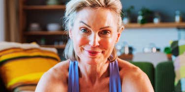People Who Are Brilliant But Constantly Surrounded By Idiots Often Say Brutally Honest Phrases A close-up portrait of a fit mature woman with blonde hair smiling directly at the camera while wearing a purple tank top during a home workout.