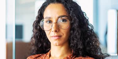 Close-up portrait of confident serious business woman, hispanic woman wearing glasses inside office looking at camera with curly hair.