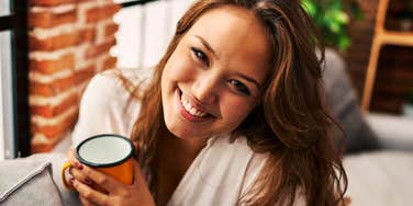 A smiling young woman leaning back on a sofa with a warm cup of coffee in her hands, looking affectionately at her partner, representing genuine happiness, daily gratitude, emotional connection, and love.
