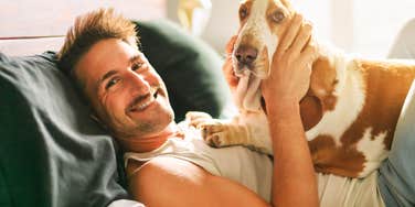 Smiling man lying on a bed with a basset hound resting on his chest in warm sunlight