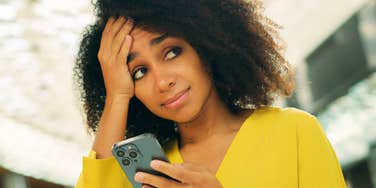 Woman with curly hair in a yellow top holding a smartphone and touching her forehead with an exasperated expression