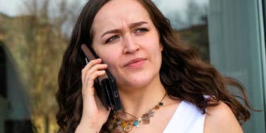 A young woman with brown wavy hair wearing a white sleeveless dress and gold charm necklace, holding a smartphone with a leather case up to her ear with a suspicious, mildly annoyed expression.