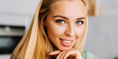 A close-up portrait of a cheerful young woman with long blonde hair and blue eyes resting her chin on her hand, smiling warmly at the camera, representing genuine happiness, wonder, and exceptional good fortune.