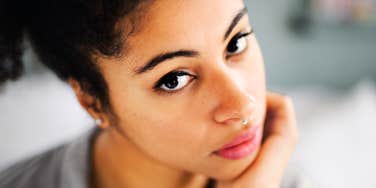 A close-up portrait of a young woman with dark curly hair tied back and a nose ring resting her cheek on her hand, looking intently into the camera with a vulnerable expression, representing emotional reflection and adult self-reliance.
