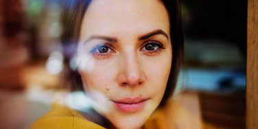 Woman with brown eyes, dark hair looking at the camera through a window with soft reflections.