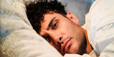 Man with dark curly hair lying on a pillow, looking sad and tired as he gazes off-camera.
